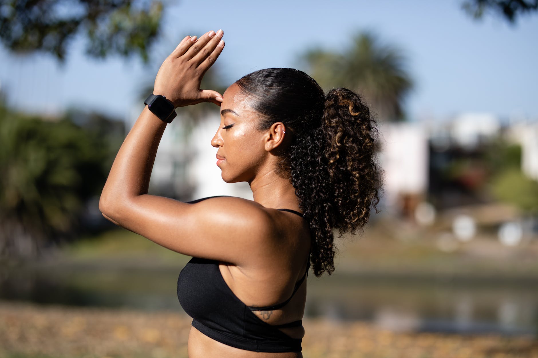 A woman practicing yoga outdoors in a serene setting, holding a mudra with her hands raised in front of her face, with closed eyes and a peaceful expression.