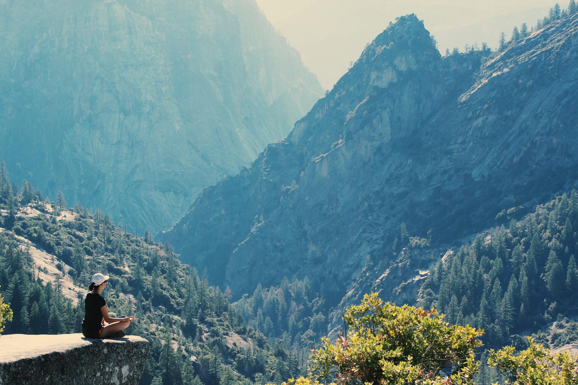 A person meditating on a rocky ledge, overlooking a breathtaking mountainous landscape with lush green trees and hazy blue peaks in the background.