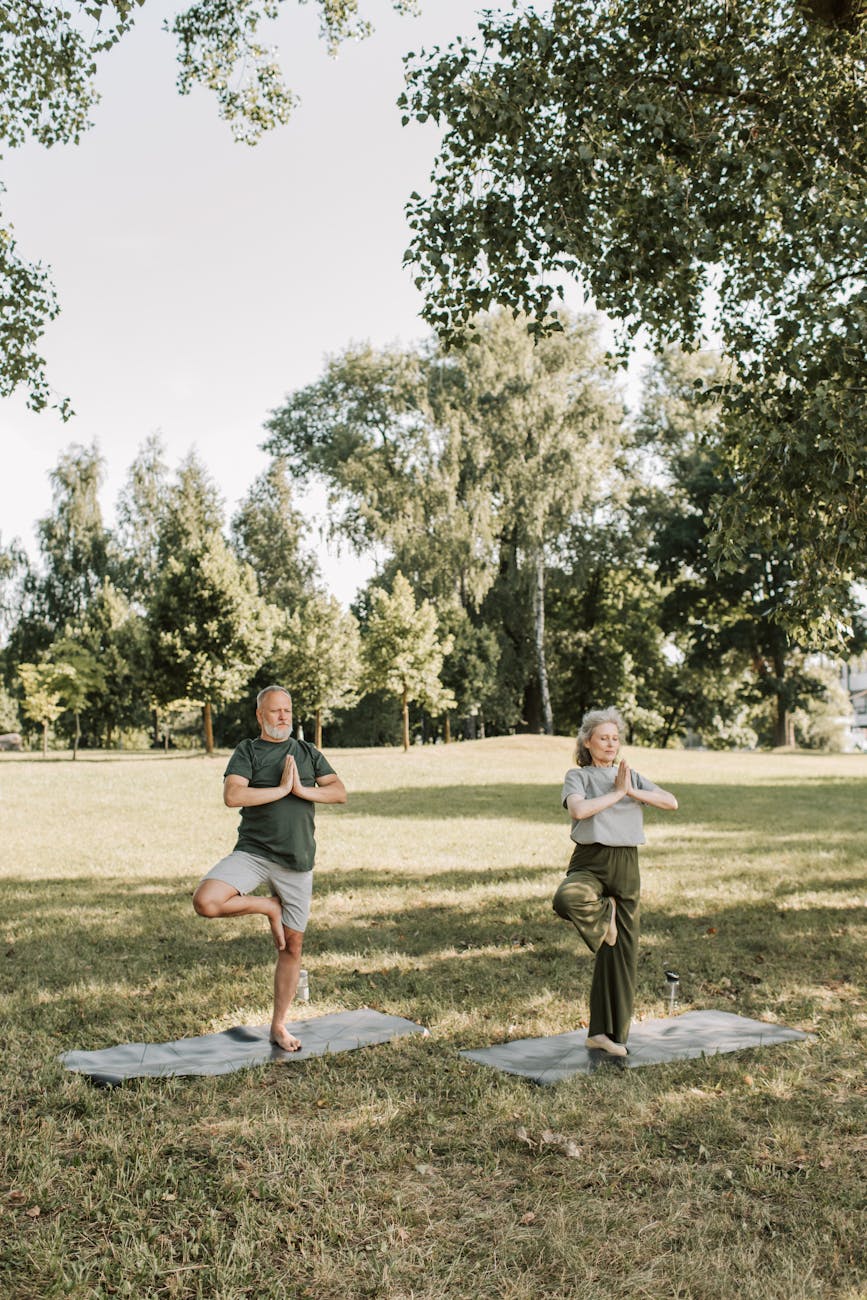 Two individuals practicing yoga outdoors on exercise mats, surrounded by trees in a grassy area.